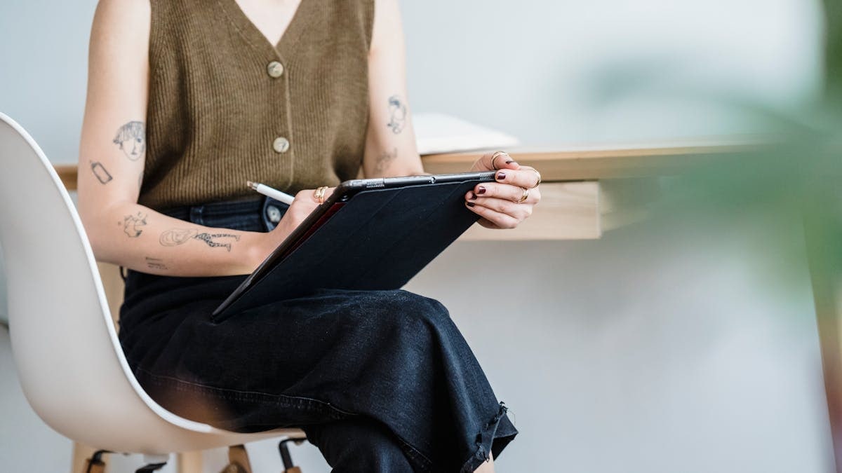 Crop unrecognizable female designer sitting on white chair and drawing in graphics tablet in selective focus
